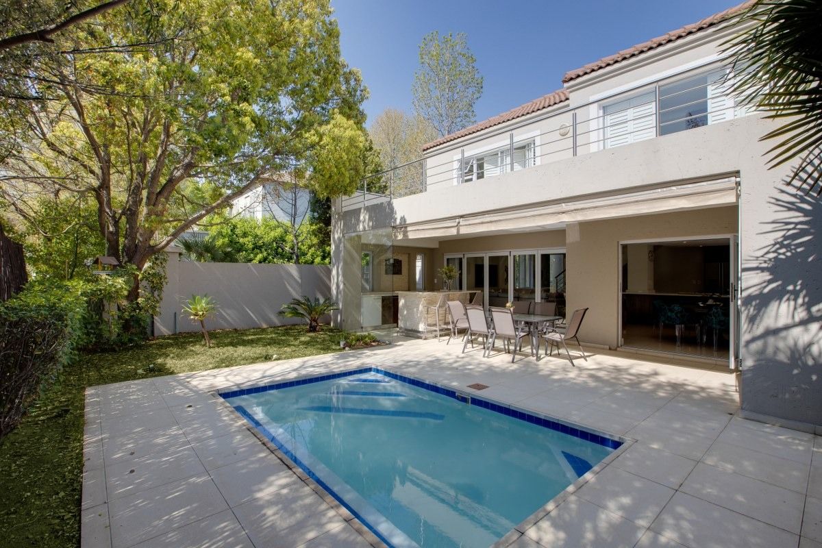 Rear exterior view featuring a swimming pool, tiled patio area, built-in bar, and an upper-level balcony overlooking the garden.