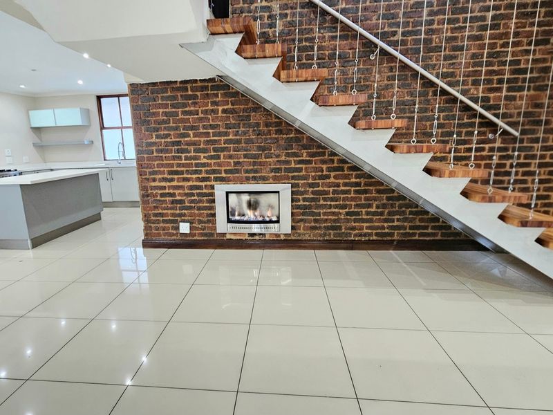Modern open-plan living area featuring a striking exposed brick wall, floating staircase, built-in gas fireplace, and a sleek kitchen with a central island.