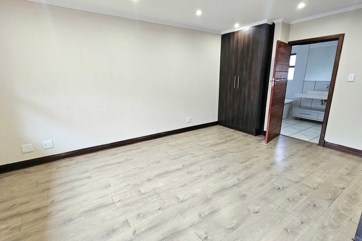 A bright, empty bedroom featuring modern laminate flooring, recessed downlighting, and a dark wood built-in wardrobe with direct access to an en-suite bathroom.
