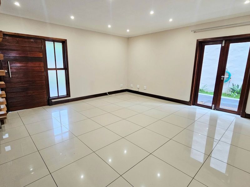 Spacious, light-filled living area featuring neutral tiled flooring, recessed lighting, and direct access to the exterior through a wooden framed glass door.