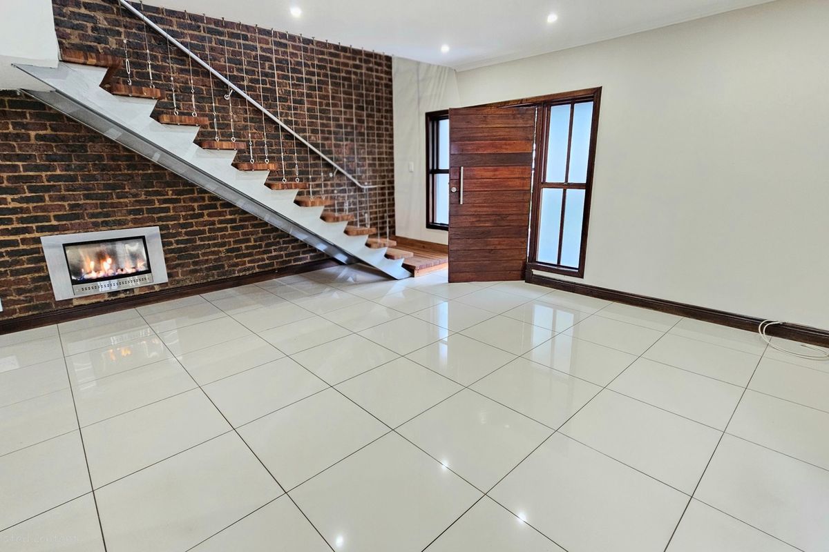 Spacious living area featuring a striking exposed brick wall, built-in fireplace, modern staircase, and polished tile flooring, creating a bright and open entrance space.