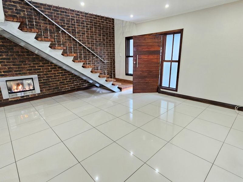 Spacious living area featuring a striking exposed brick wall, built-in fireplace, modern staircase, and polished tile flooring, creating a bright and open entrance space.