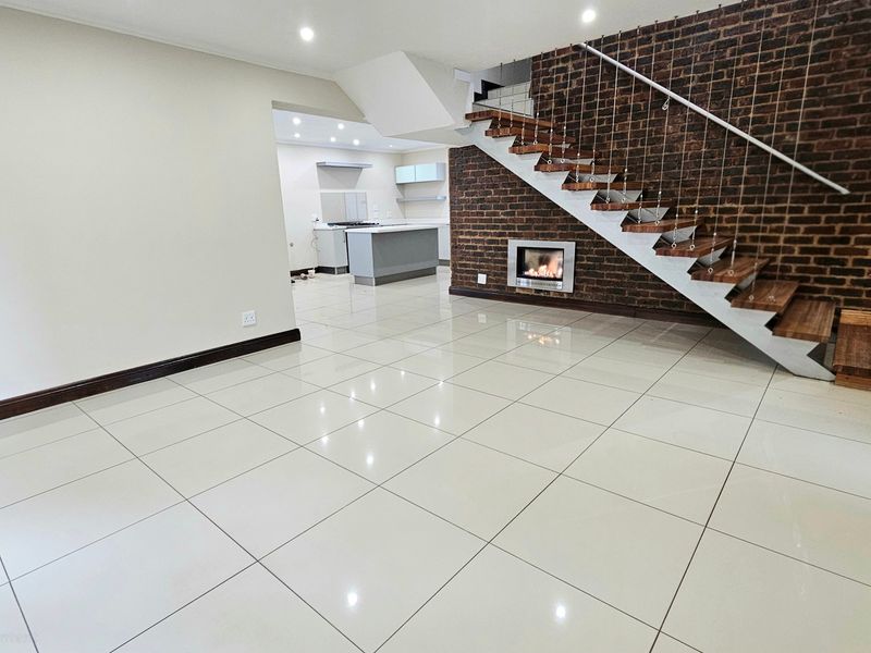 Spacious open-plan living area featuring polished tile floors, a striking exposed brick wall with a built-in fireplace, and a modern staircase leading to the upper level.