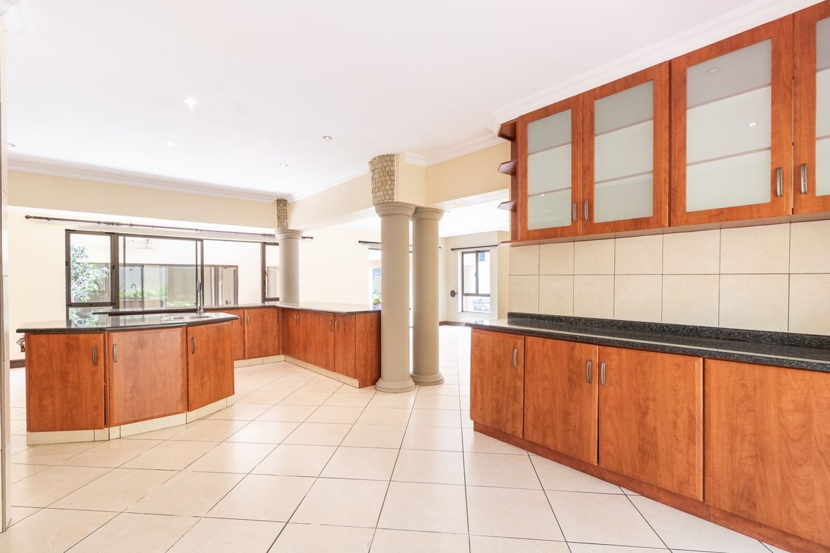 Spacious open-plan kitchen featuring warm-toned wooden cabinetry, granite countertops, and neutral ceramic floor tiles, complemented by decorative columns and recessed lighting.