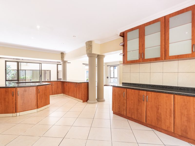 Spacious open-plan kitchen featuring warm-toned wooden cabinetry, granite countertops, and neutral ceramic floor tiles, complemented by decorative columns and recessed lighting.