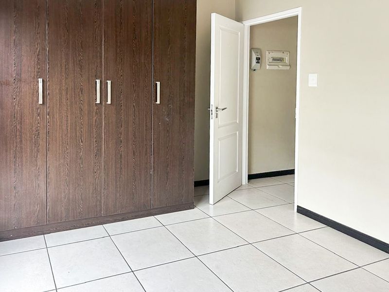 A clean, empty bedroom featuring dark wood built-in cupboards, neutral tiled flooring, and a doorway leading to an adjacent hallway area.