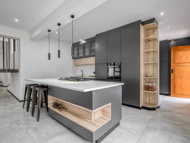 Modern kitchen featuring a central island with gas hob, sleek dark cabinetry, integrated oven, and light wood shelving accents on polished tiled floors.