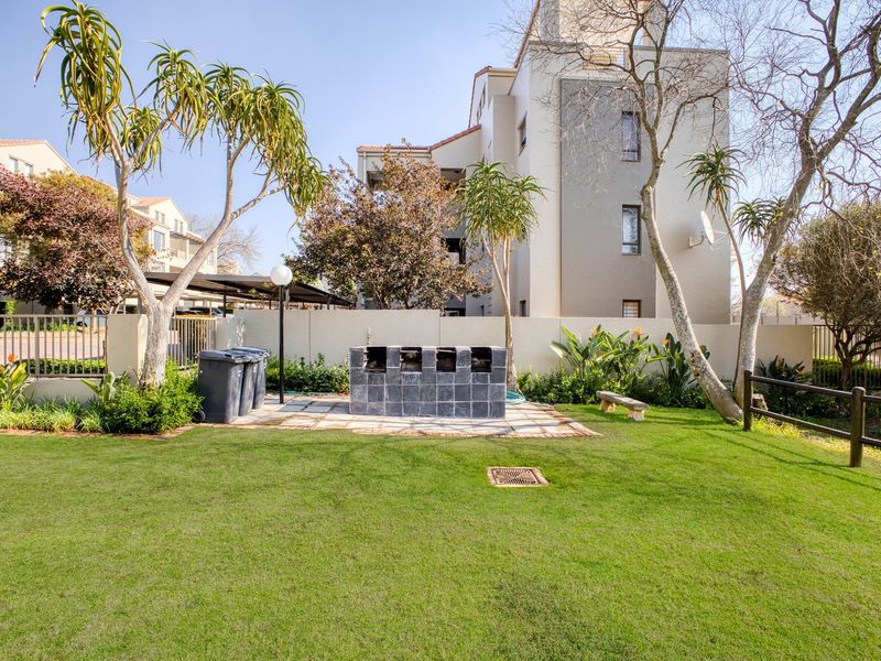 A well-maintained communal garden area featuring a built-in brick braai, paved patio section, and established landscaping within an apartment complex.