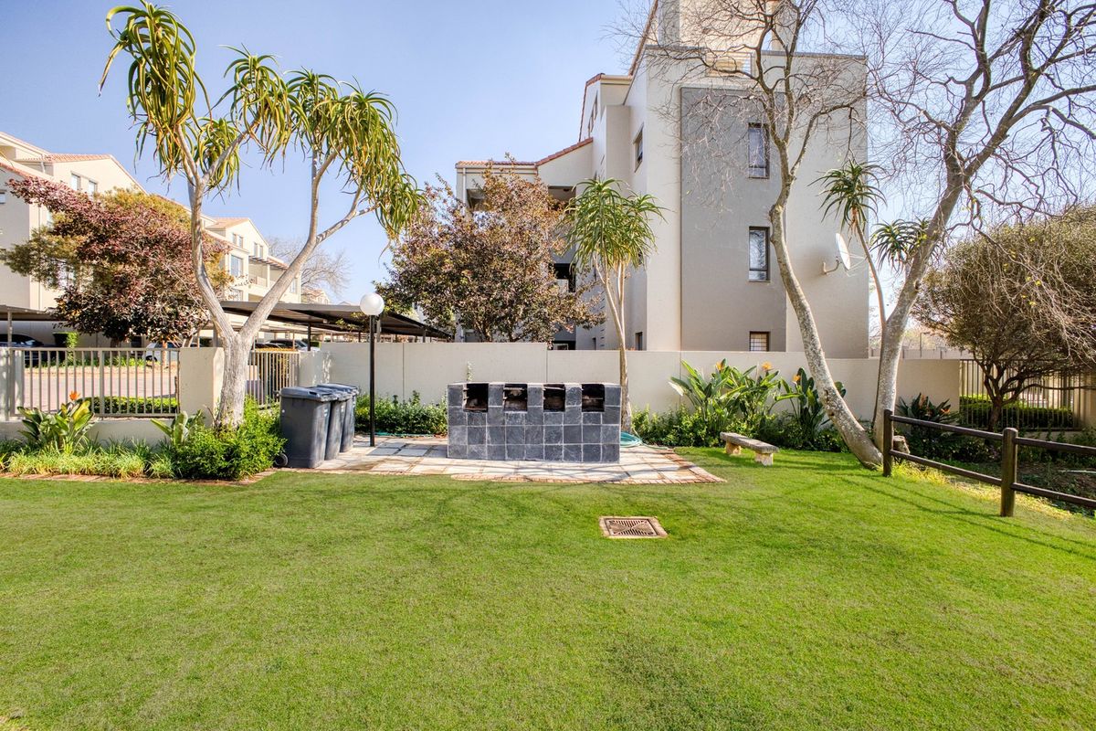 A well-maintained communal garden area featuring a built-in brick braai, paved patio section, and established landscaping within an apartment complex.