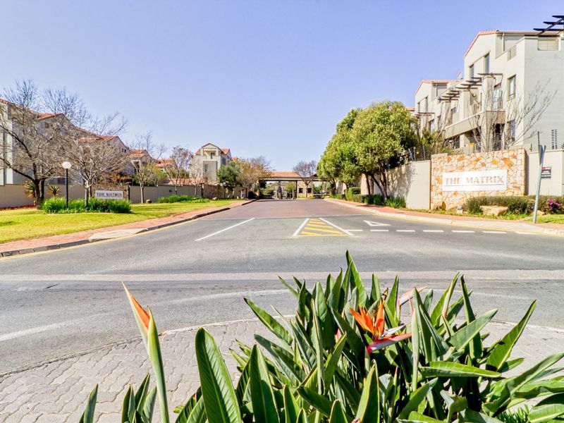 Street view of The Matrix residential complex entrance, featuring paved access road, security gatehouse, and well-maintained landscaped surroundings.