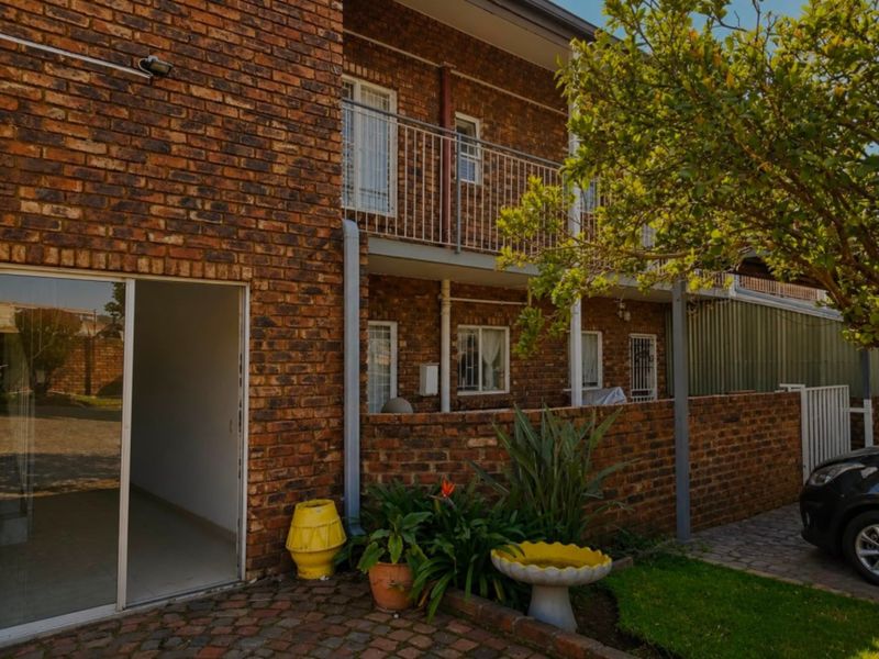Rear exterior view of a brick apartment building featuring a paved patio area, sliding glass door access, and an upper-level balcony with metal railings.