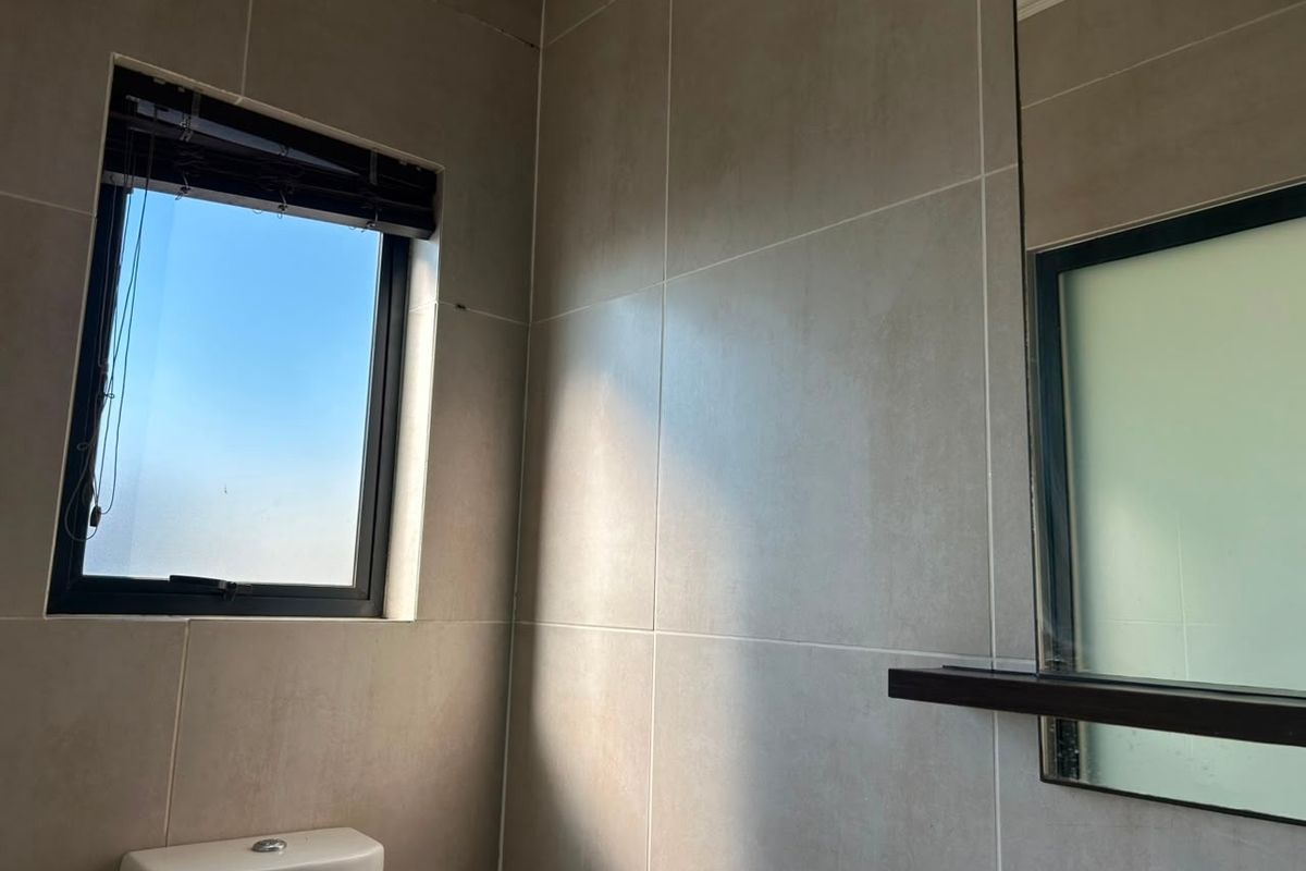 A modern bathroom featuring neutral wall tiles, a wall-mounted mirror with a shelf, and a window providing natural light.