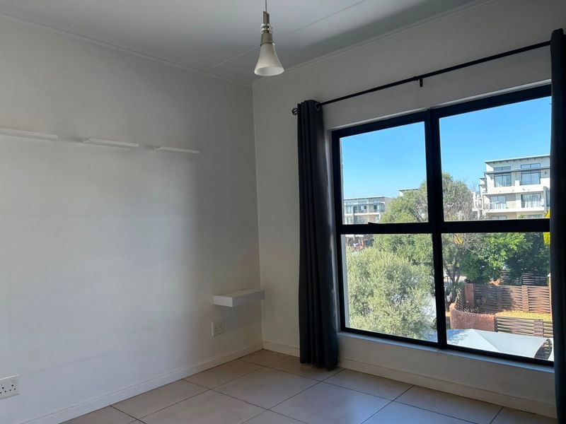 A bright, empty bedroom featuring tiled flooring, a large window with neighbourhood views, and minimalist wall-mounted shelving.