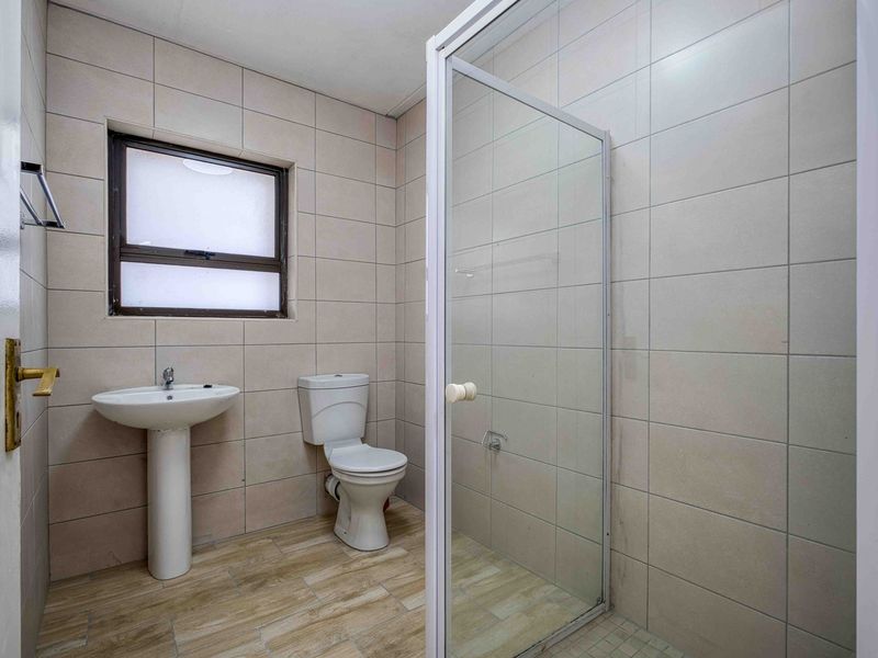 A clean, functional bathroom featuring a pedestal basin, toilet, and a glass-enclosed shower with neutral wall and floor tiling.