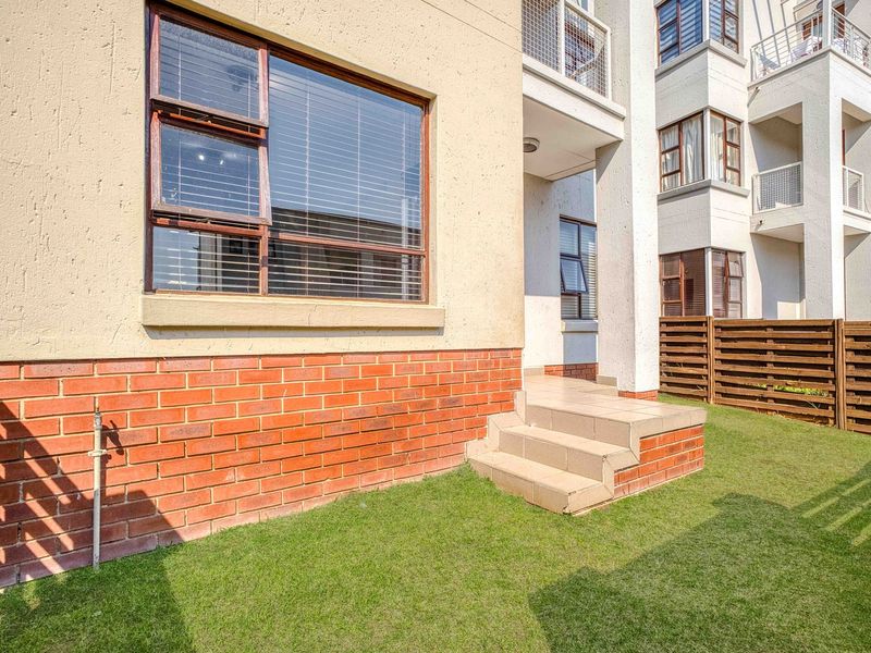 A view of the rear exterior of the apartment building, featuring a small private garden area, brick base detailing, and tiled entrance steps.