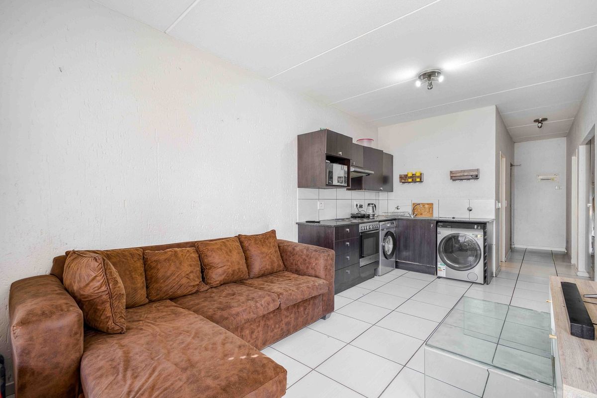 Open-plan living area featuring a compact kitchen with built-in cabinetry, electric stove, and integrated laundry facilities, finished with neutral floor tiles.