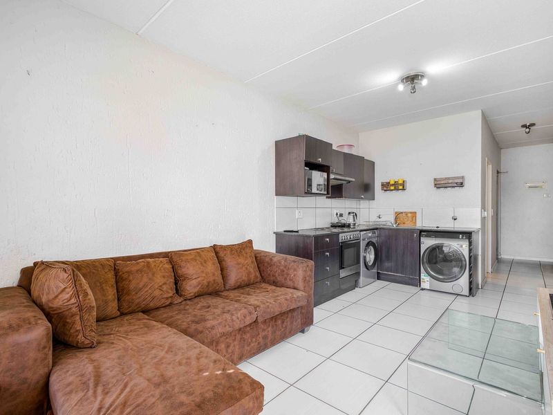Open-plan living area featuring a compact kitchen with built-in cabinetry, electric stove, and integrated laundry facilities, finished with neutral floor tiles.