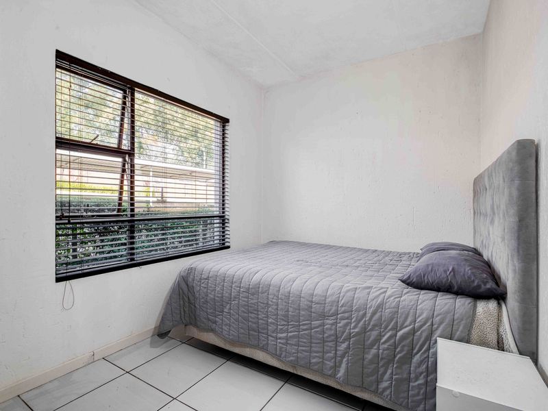 A compact bedroom featuring tiled flooring, a large window with venetian blinds, and neutral wall finishes.