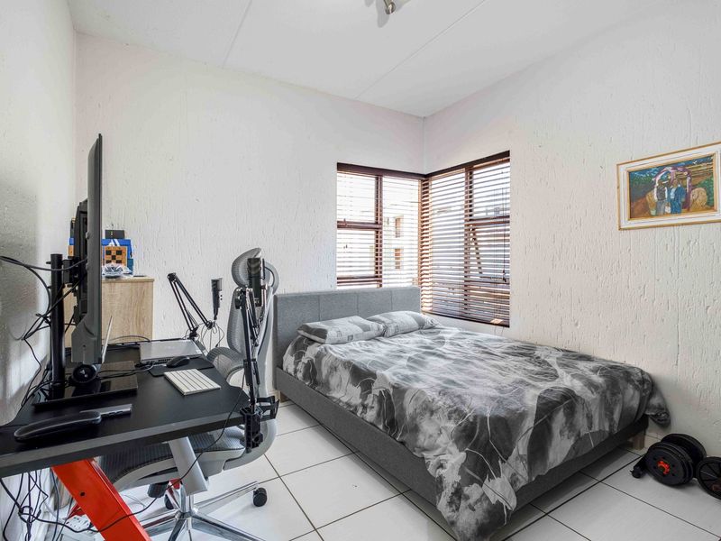A functional bedroom featuring tiled flooring and a corner window with fitted venetian blinds, providing natural light and a compact layout.
