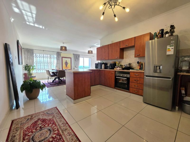 Open-plan living and kitchen area featuring tiled flooring, wooden cabinetry, and integrated appliances, with access to a balcony through a security gate.
