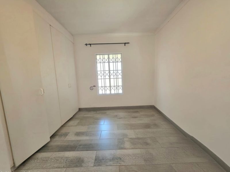 A clean, empty bedroom featuring neutral tiled flooring, built-in cupboards for storage, and a window fitted with a security gate.