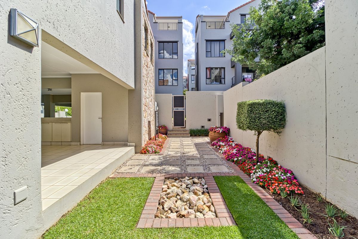 A well-maintained rear courtyard featuring a paved patio, landscaped garden beds, a decorative stone feature, and secure boundary walls.