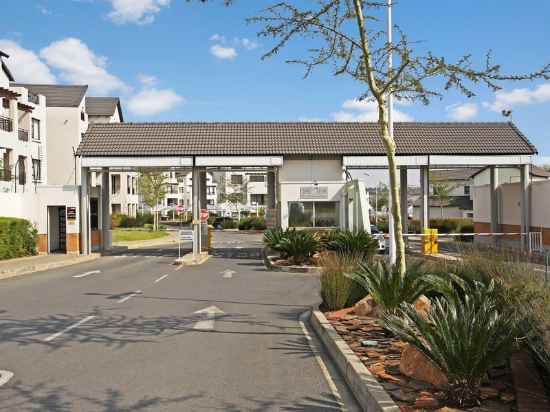 The secure entrance gatehouse of the apartment complex, featuring a boom gate, paved driveway, and well-maintained landscaped surroundings.