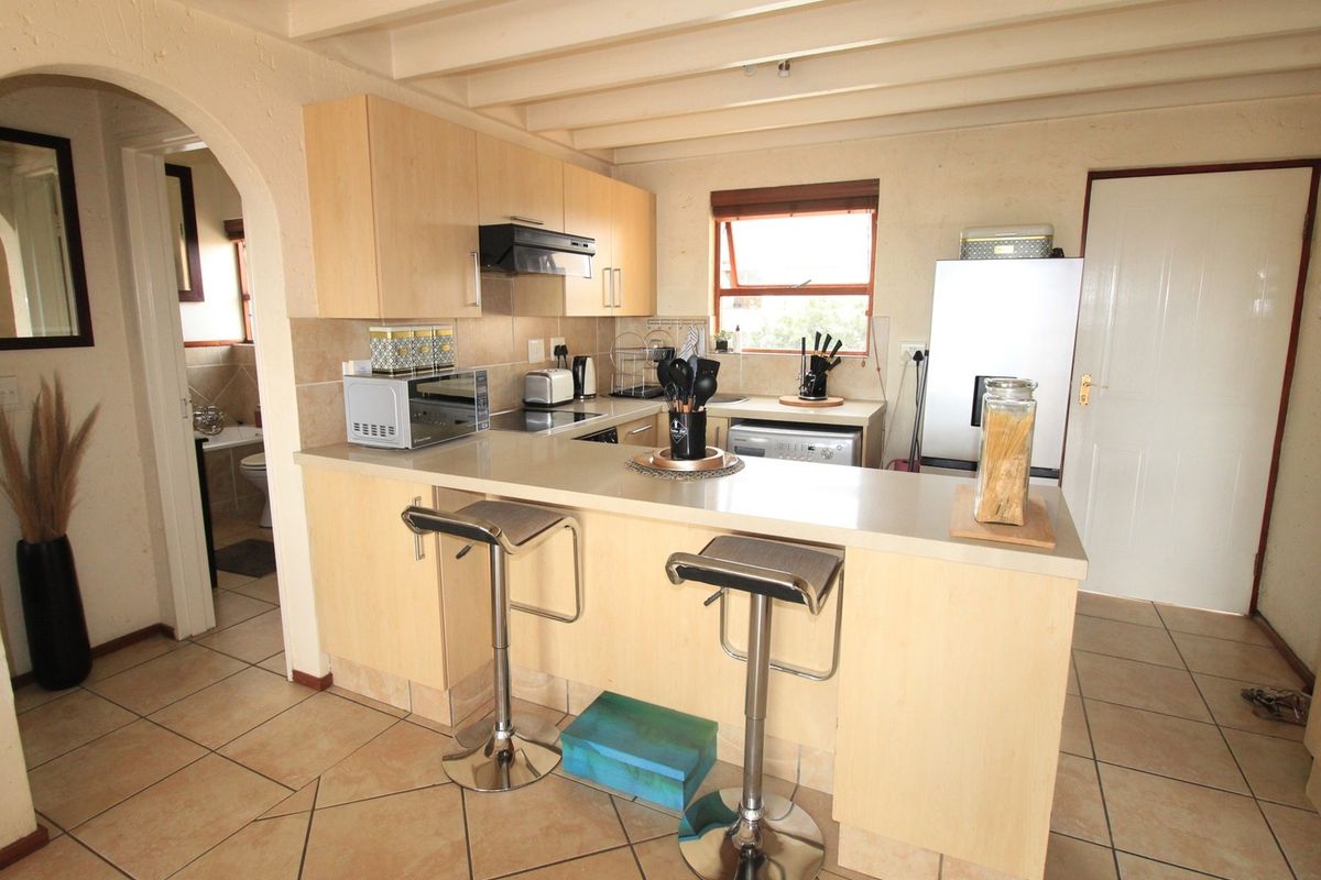 A functional kitchen featuring light wood-finish cabinetry, a breakfast bar, tiled flooring, and an exposed beam ceiling, with an adjacent archway leading to the bathroom.