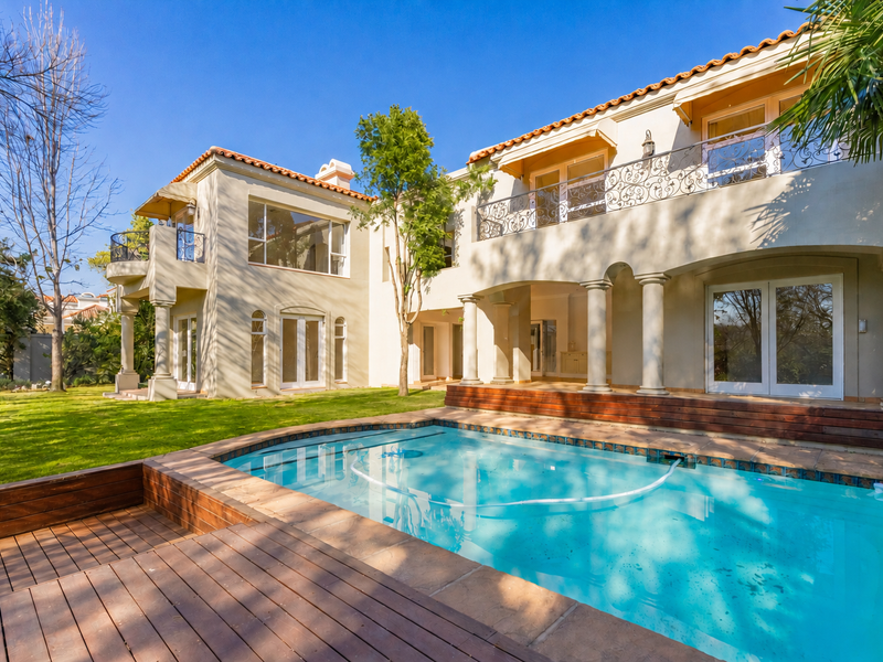 Rear elevation of a two-story residence featuring a swimming pool, wooden decking, and a balcony with decorative wrought iron railings.
