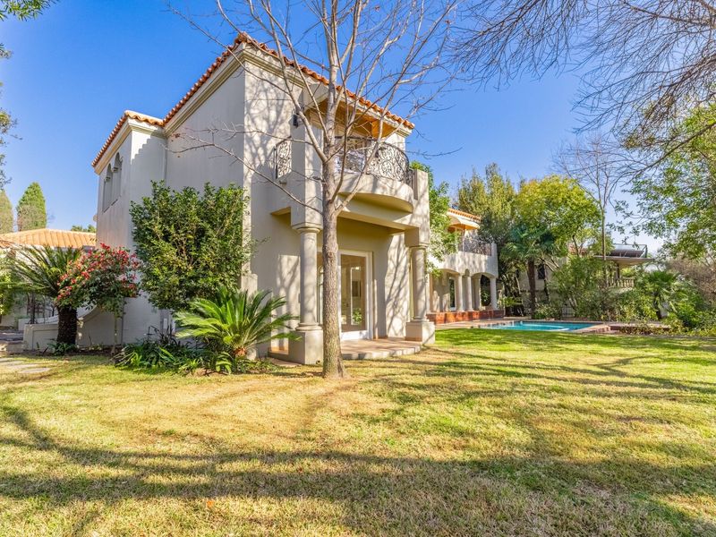 Rear view of the property featuring a landscaped garden, swimming pool, and a balcony with wrought iron railings and classic architectural columns.