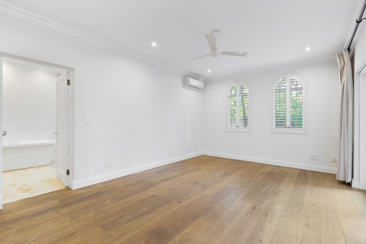 Spacious bedroom featuring hardwood flooring, arched windows with plantation shutters, a ceiling fan, and an en-suite bathroom.