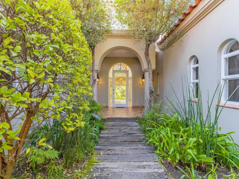 A charming arched entryway featuring tiled flooring, classic arched windows, and a rustic wooden walkway surrounded by lush garden landscaping.