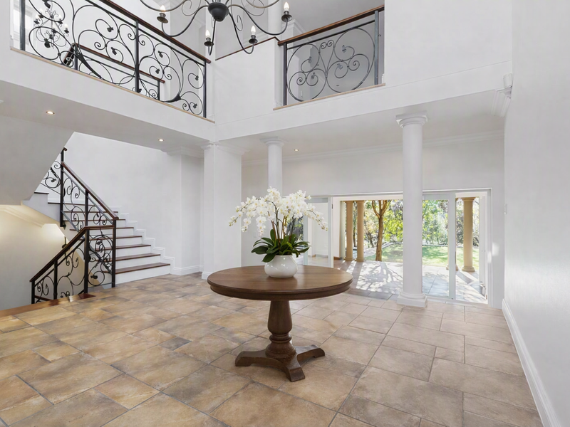 Grand double-volume entrance hall featuring stone tile flooring, a sweeping staircase with wrought iron balustrades, and classical columns leading to a rear patio.