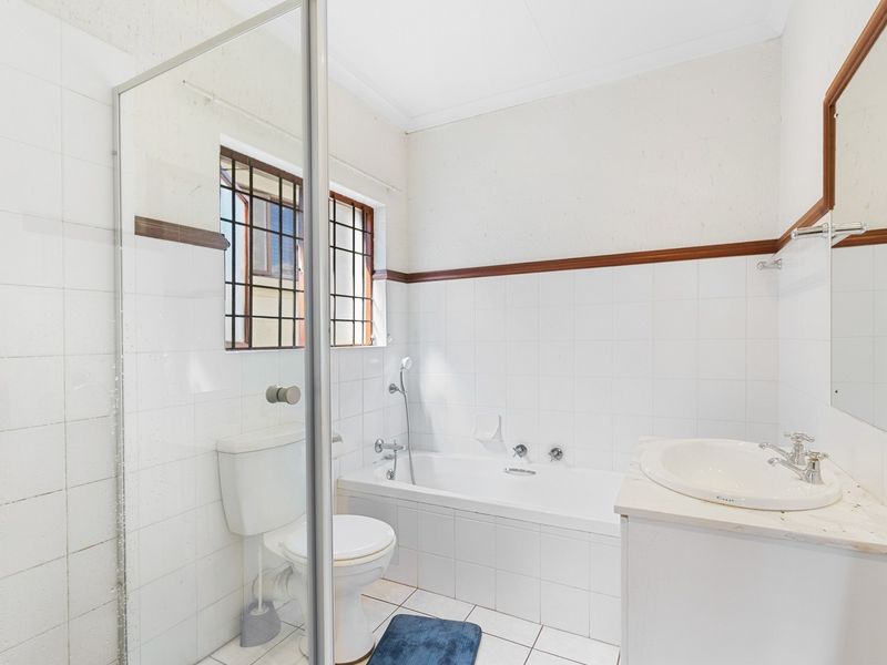 A functional bathroom featuring a built-in bathtub, glass shower screen, vanity basin, and toilet with white tiled finishes and security window bars.