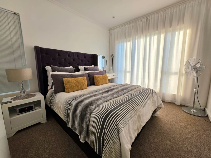 A well-lit bedroom featuring carpeted flooring, neutral walls, and large windows with floor-to-ceiling curtains, providing a comfortable and bright living space.