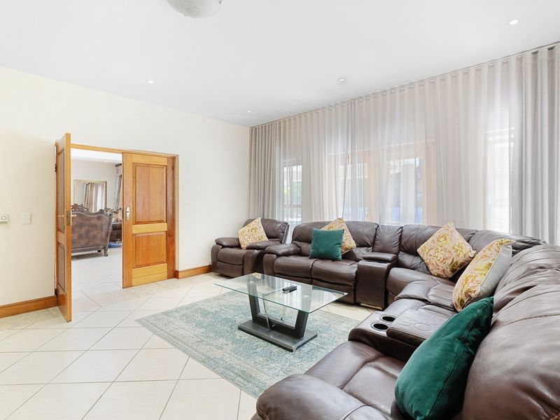 A spacious living room featuring tiled flooring, recessed downlights, and large windows dressed with sheer curtains, providing access to an adjacent room.