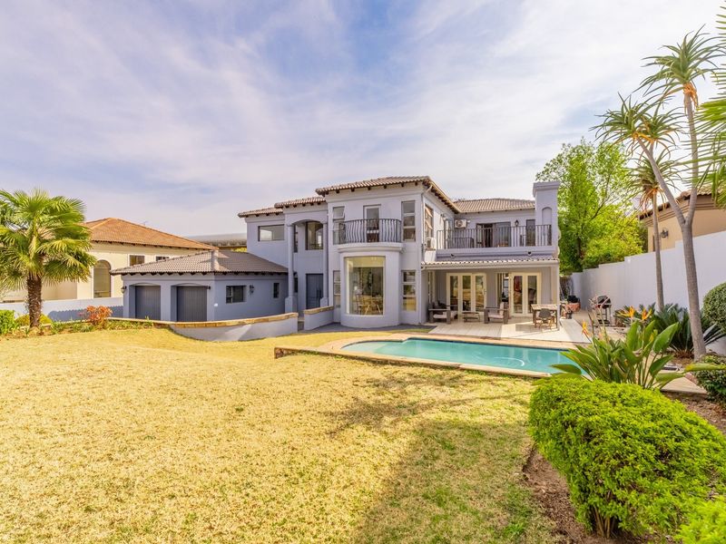 Rear view of a two-story house featuring a swimming pool, paved patio area, upper-level balcony, and a well-maintained garden with a double garage.