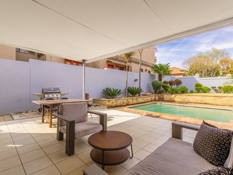 A covered patio area featuring tiled flooring, overlooking a private swimming pool with a landscaped retaining wall and garden backdrop.