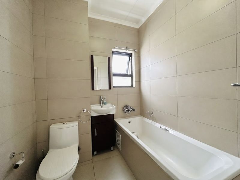 A clean, modern bathroom featuring neutral wall tiles, a built-in bathtub, a vanity basin, and a toilet.