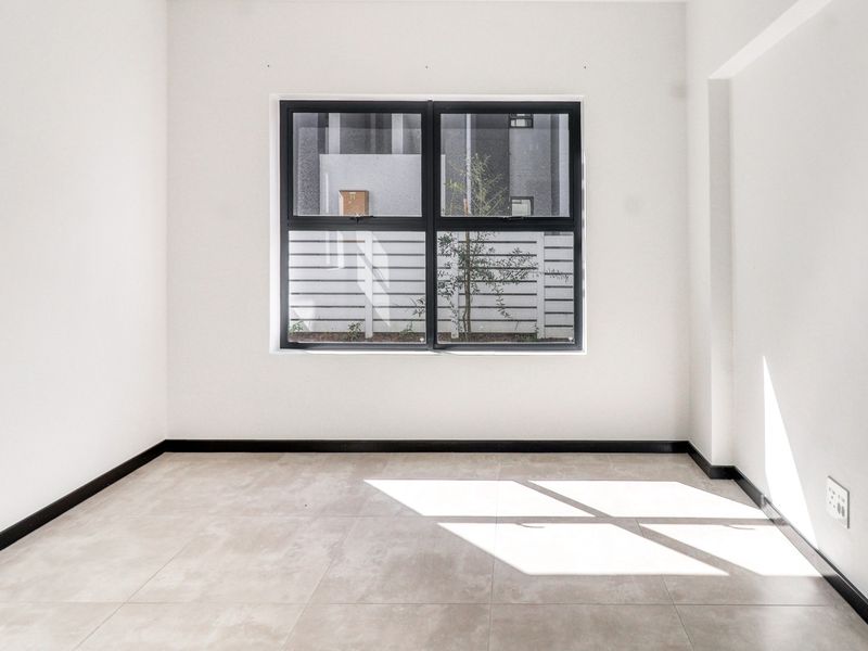 A bright, empty bedroom featuring modern tiled flooring, clean white walls, and a large black-framed window providing natural light.