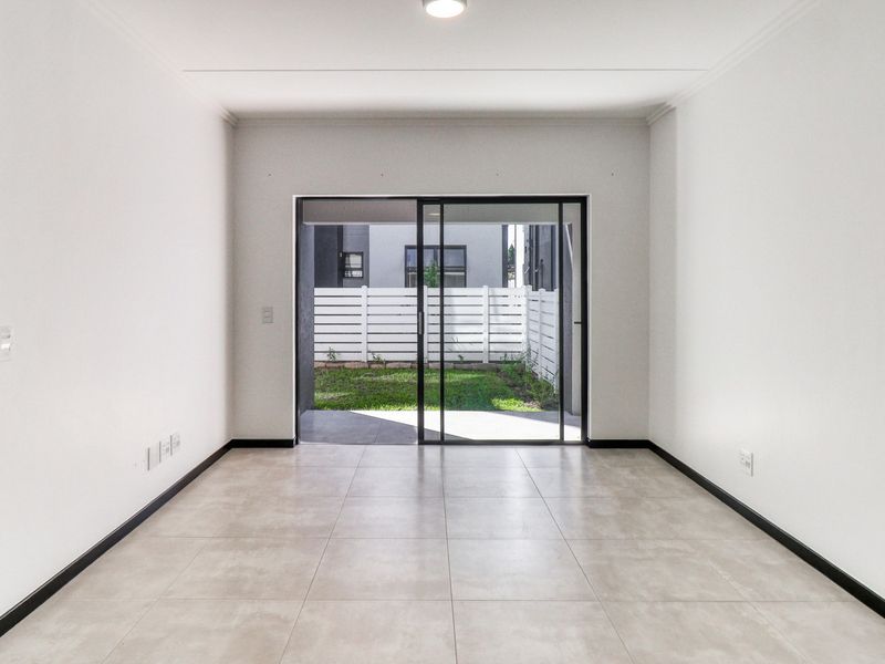 A bright, empty living area featuring neutral tiled flooring and a large sliding glass door providing direct access to the outdoor patio and garden.