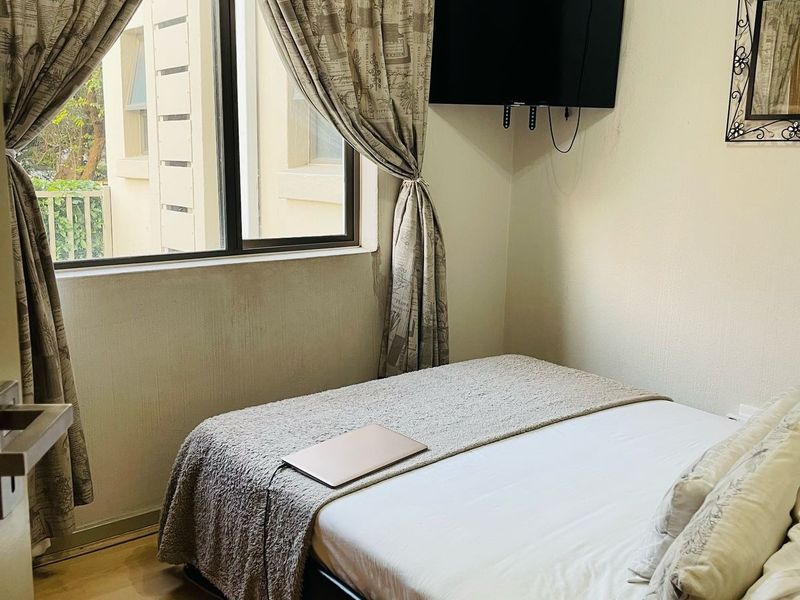 A compact bedroom featuring laminate flooring, a window with curtains, and a wall-mounted television bracket.