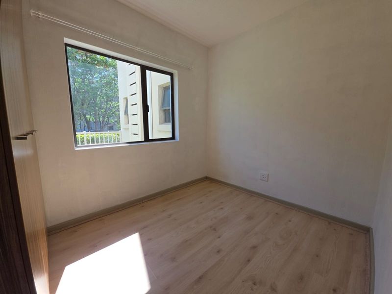 A bright, empty bedroom featuring light-toned laminate flooring, a built-in cupboard, and a large window providing natural light.