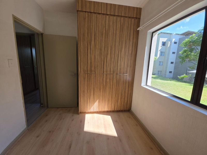 A bright, empty bedroom featuring modern wood-grain built-in wardrobes, light laminate flooring, and a large window overlooking the exterior grounds.