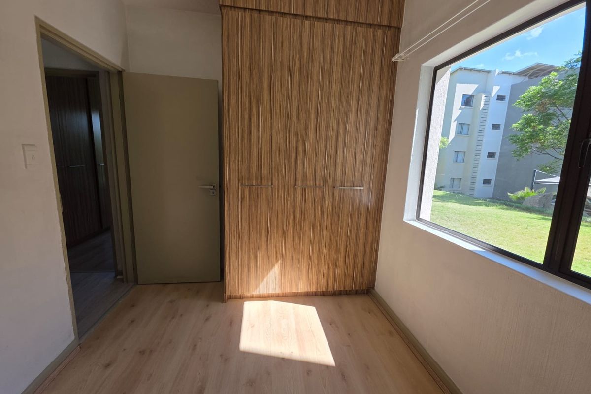 A bright, empty bedroom featuring modern wood-grain built-in wardrobes, light laminate flooring, and a large window overlooking the exterior grounds.