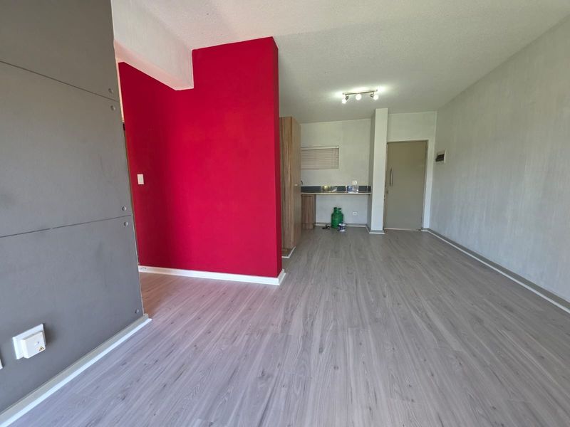 An open-plan living and kitchen area featuring light-toned laminate flooring, a bold red accent wall, and basic built-in kitchen cabinetry.