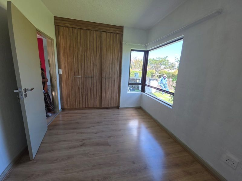 A bright, empty bedroom featuring modern laminate flooring, built-in wooden cupboards, and a large corner window providing natural light.