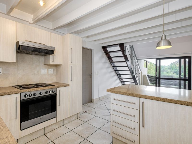 Modern open-plan kitchen featuring light wood cabinetry, stone countertops, and integrated appliances, flowing into a living area with a staircase and access to the outdoors.