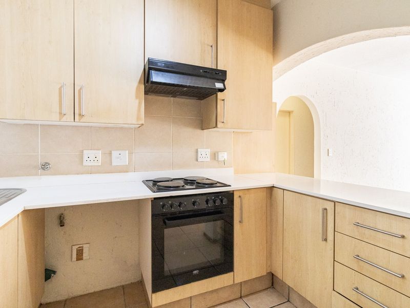 A functional kitchen featuring light wood-finish cabinetry, a built-in oven with hob, extractor fan, and tiled backsplash, leading into an arched hallway.