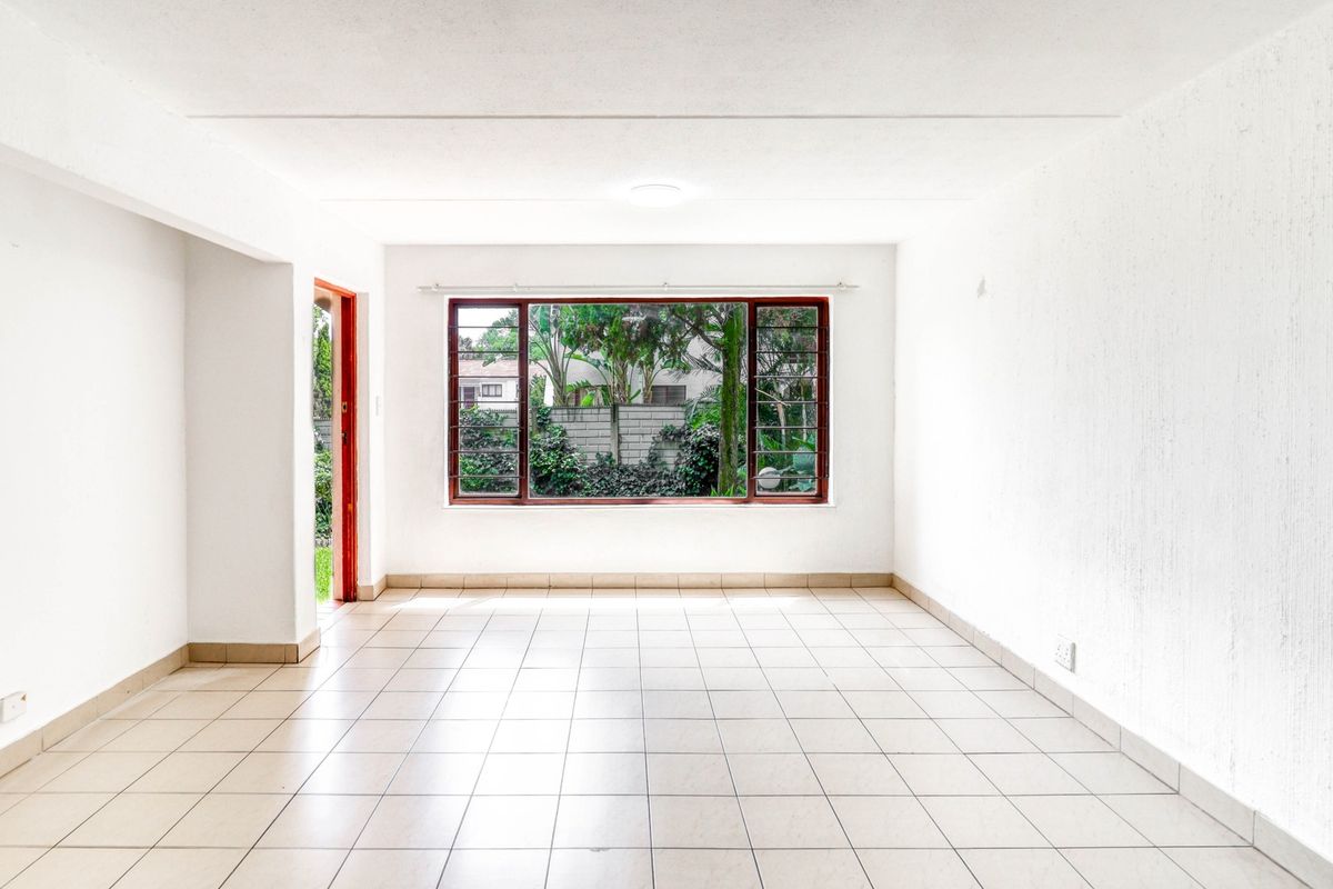 A bright, empty living room featuring neutral tiled flooring, large windows with security bars, and direct access to an outdoor area.